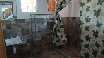 Une salle de bain avec un sol carrelé, un lavabo, une étagère en métal et un rideau de douche orné d’un motif de feuilles.