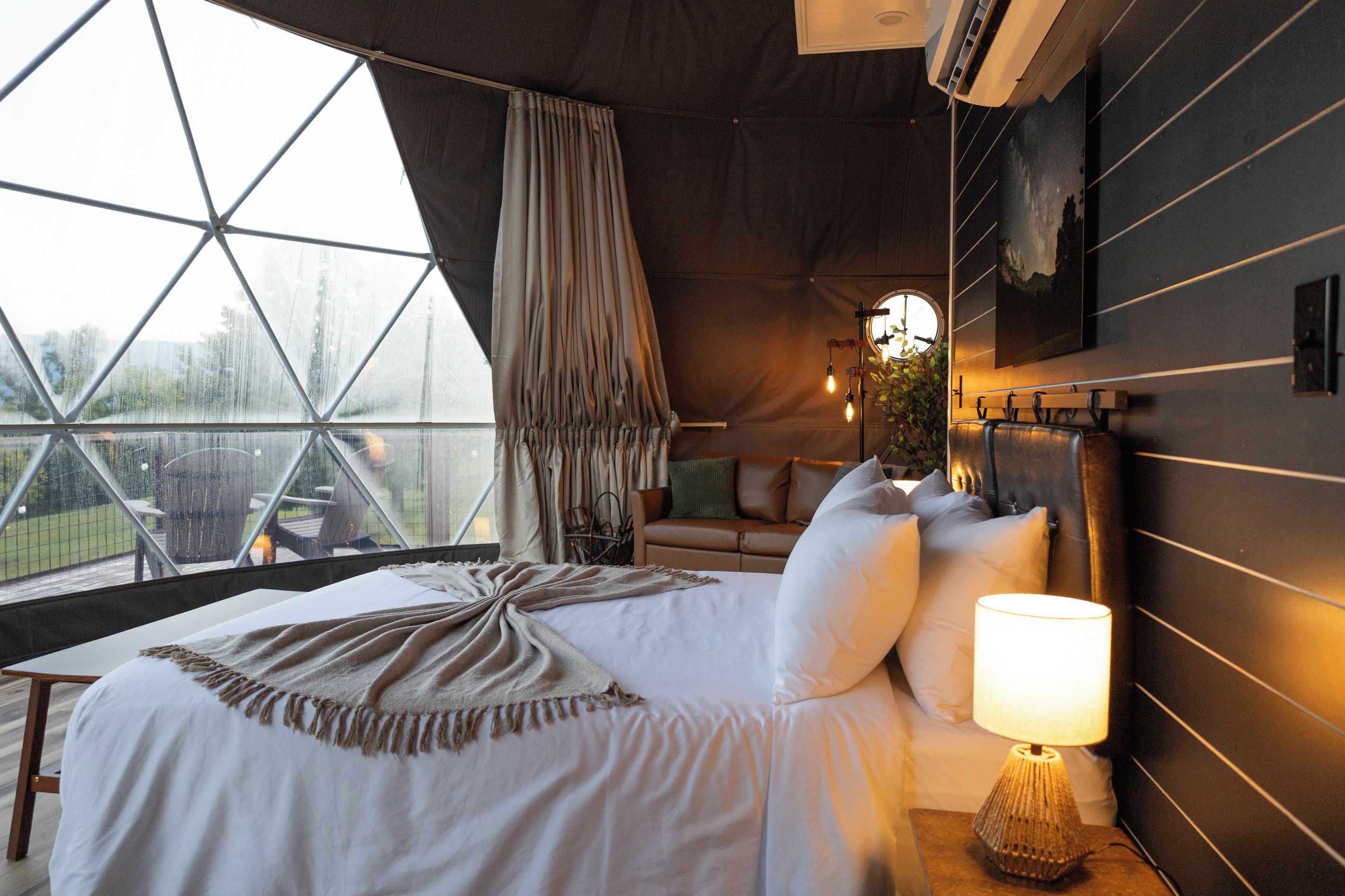 A bedroom with a large window, a bed with white bedding, a nightstand with a lamp, and a view of the outdoors.