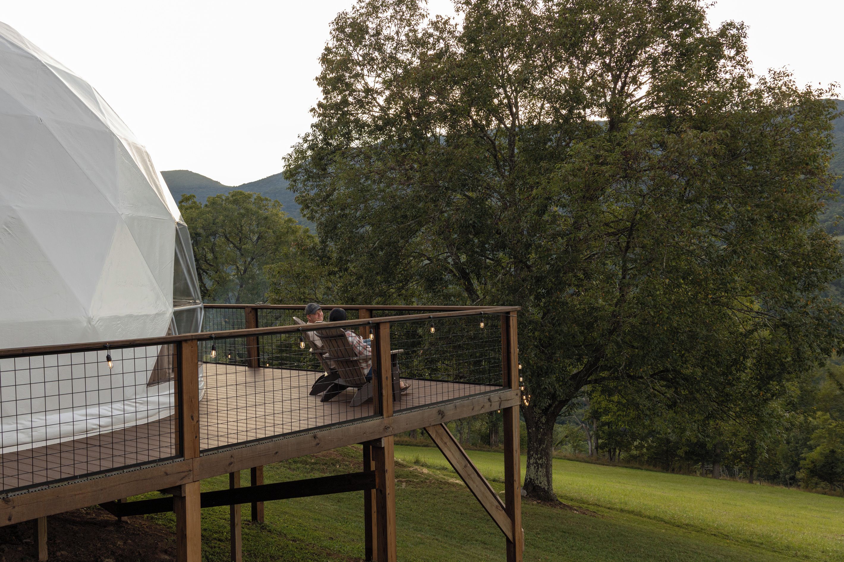 A geodesic dome structure with a wooden deck and chairs overlooking a grassy area with trees.