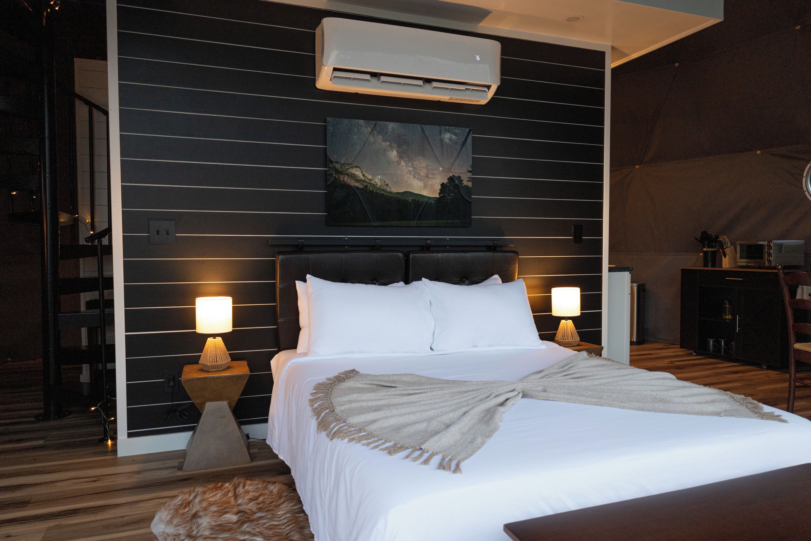 A hotel room with a bed, two bedside lamps, an air conditioning unit, a painting on the wall, and a small table with a lamp.