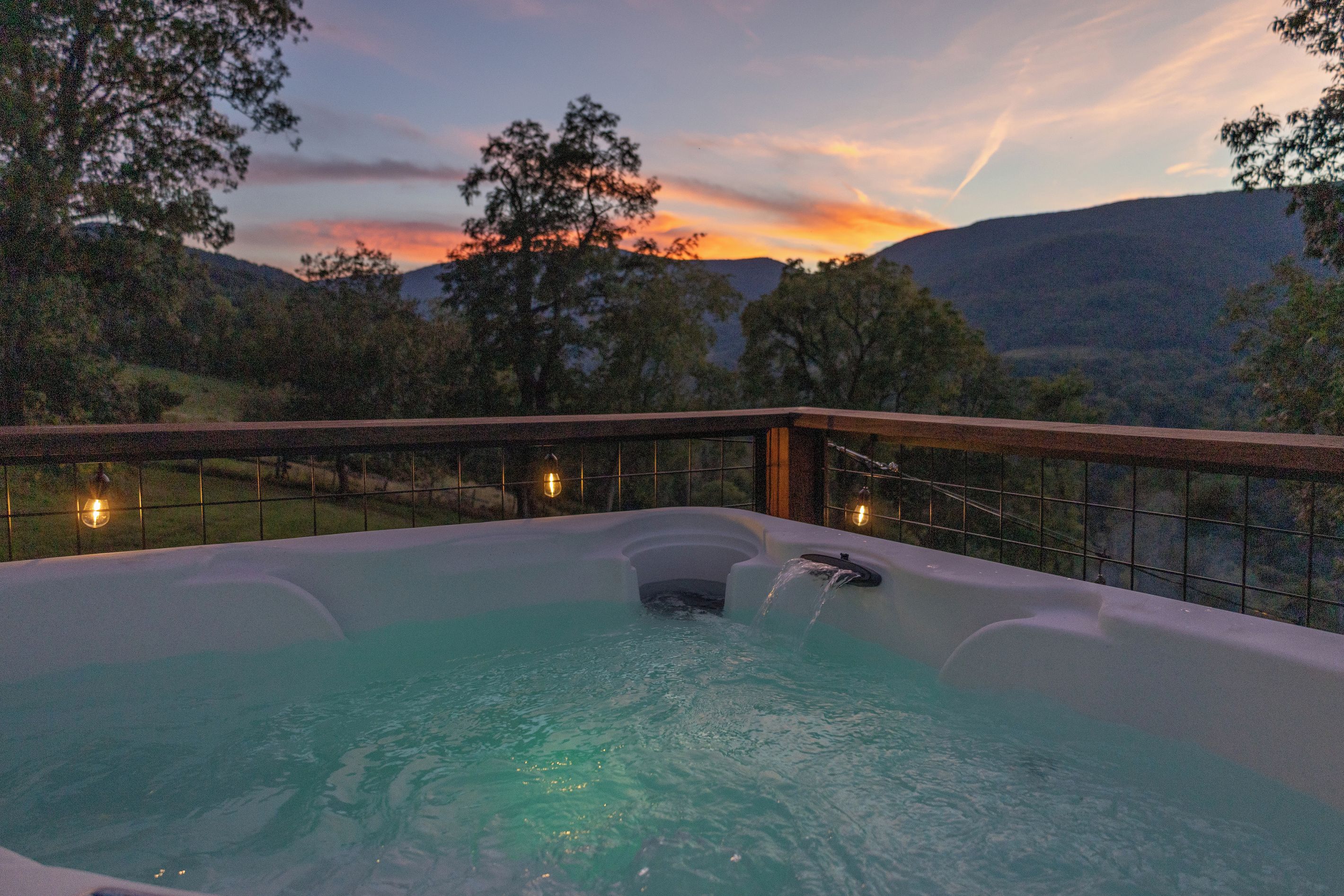 A hot tub with a wooden deck and a view of the sunset and hills.