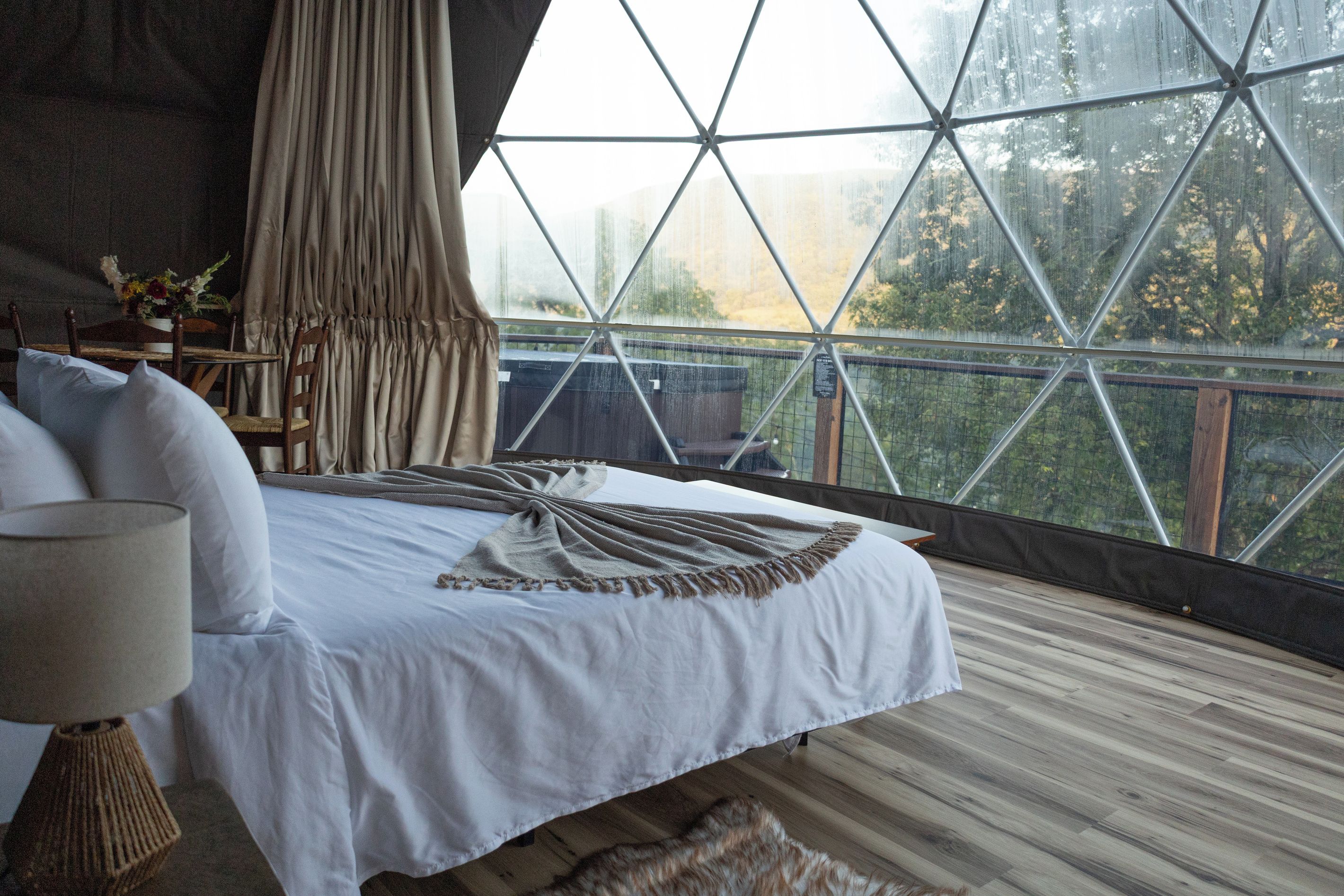 A bedroom with a large window, a bed, a nightstand, and a view of the outdoors.