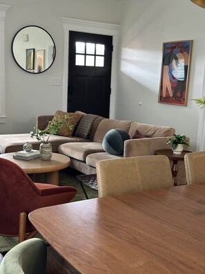 A living room with a sofa, a round coffee table, a dining table with chairs, a black door, a round mirror, and a framed painting on the wall. - Casa Vida on the Plaza (Kansas City)