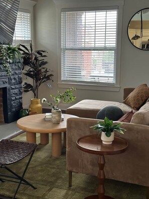 A living room with a sofa, a coffee table, and a fireplace. There are plants on the coffee table and a window with blinds. - Casa Vida on the Plaza (Kansas City)