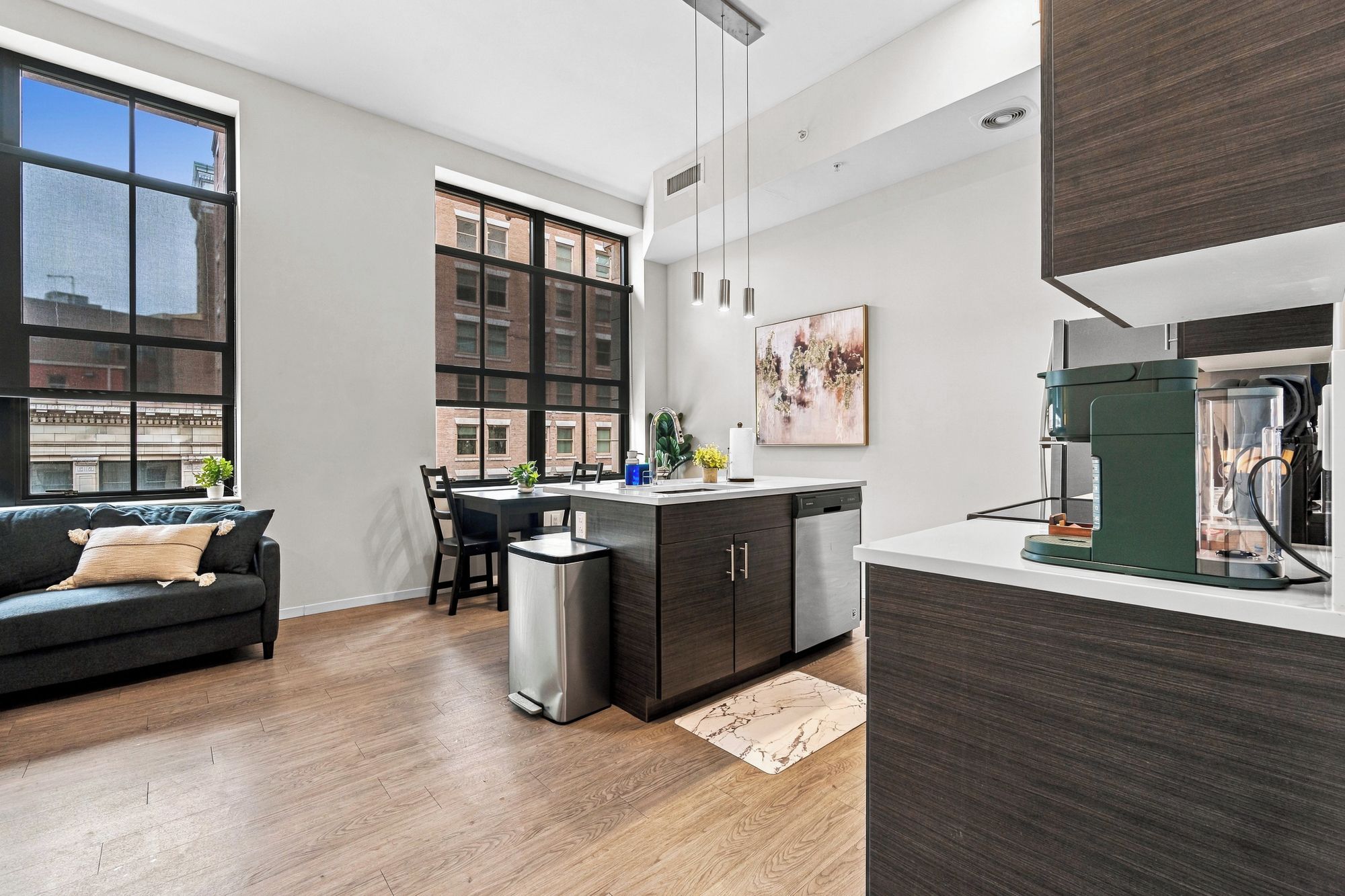 A modern kitchen with a built-in coffee machine, a dining area with a sofa and chairs, and large windows offering a city view.