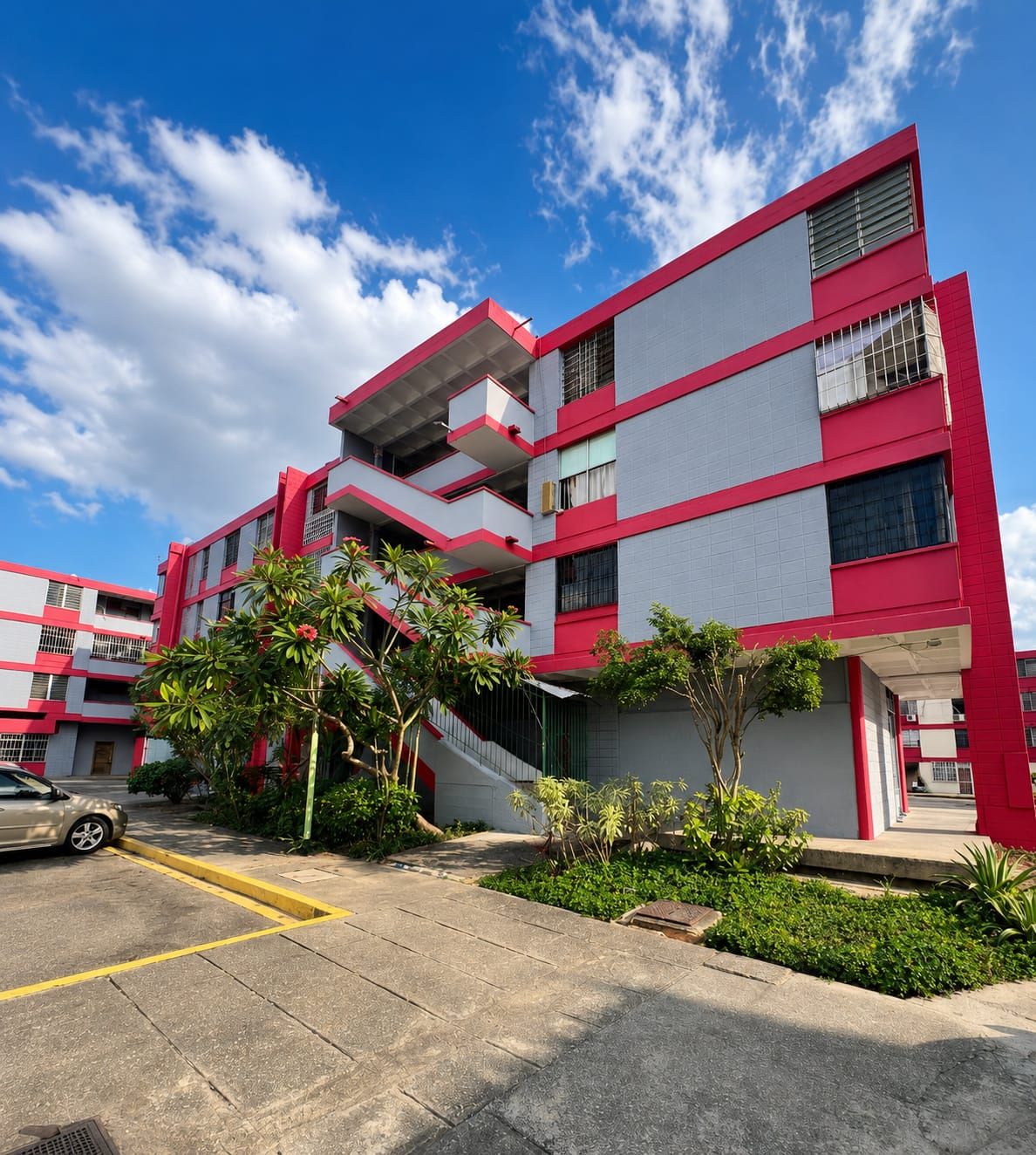 Un edificio de varios pisos con fachada roja y gris, un estacionamiento y algunas zonas verdes.