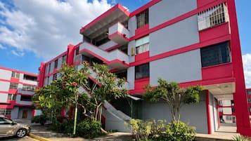 Un edificio de varios pisos con fachada roja y gris, un estacionamiento y algunas zonas verdes.