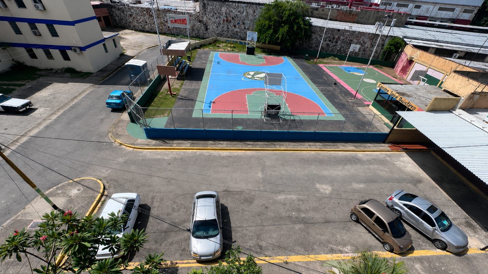 An outdoor basketball court with a blue and red surface, surrounded by a low wall and a parking area with cars.