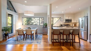 A modern kitchen with a central island, a dining area with chairs, and a large window providing natural light. - New Listing | Beautifully Updated 3BR | Close to Village & Lost Lake | Managed By Aloha Whistler (Whistler)
