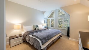 Ein Schlafzimmer mit Bett, Nachttischen, einer Lampe, einem Fenster mit Jalousien und Blick auf Bäume.
