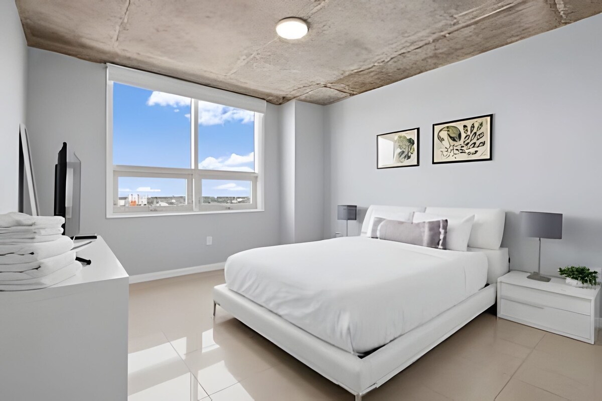 A modern bedroom with a large bed, a desk with a computer, and a window with a city view.