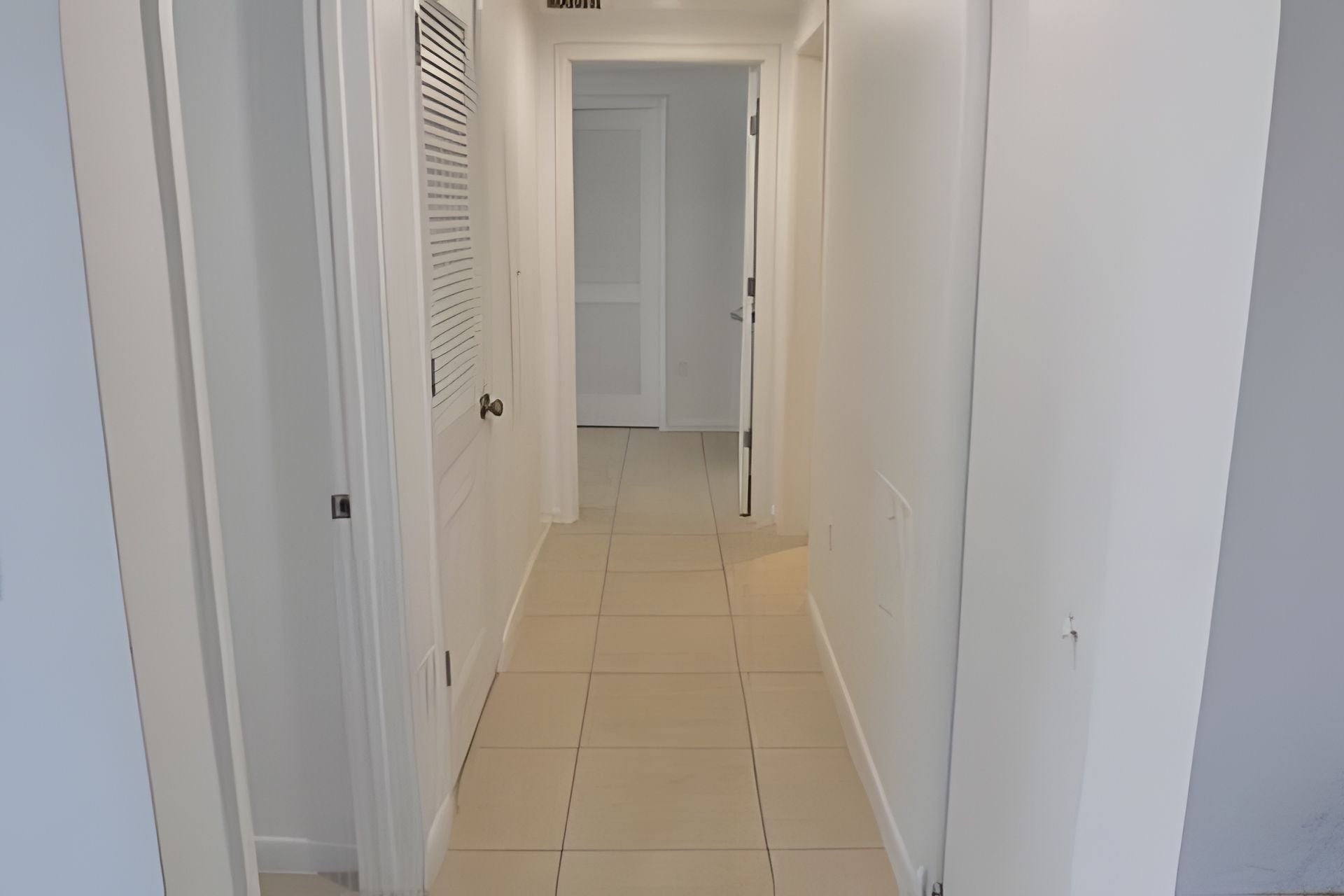 A narrow hallway with white walls and a tiled floor, leading to a door at the end.