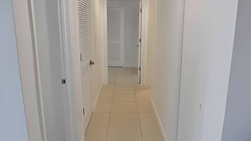 A narrow hallway with white walls and a tiled floor, leading to a door at the end.