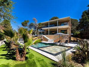 Et moderne hus i to etager med swimmingpool, udendørs møbler og frodig beplantning.