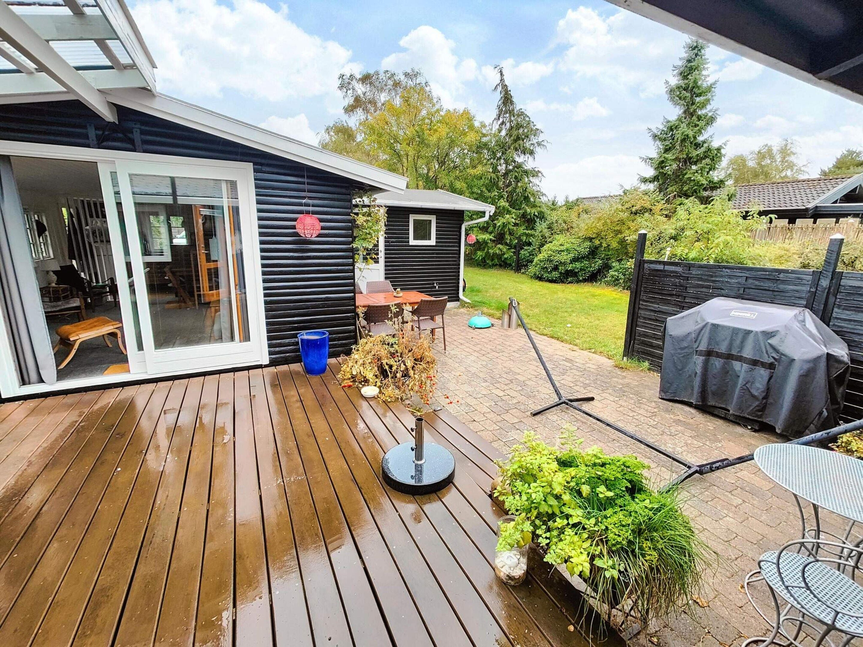En træterrasse med et udendørs bord og stole, et lille haveareal med planter, og en overdækket grill.