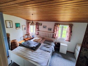 Ein Schlafzimmer mit Holzbalkendecke, einem Bett mit Kissen und Decken, einem Sessel, einem Nachttisch, einer Lampe und einem Fenster mit Vorhängen.