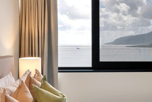 Una camera d'albergo con un letto, cuscini, una lampada e una finestra vista mare e un'isola all'orizzonte.