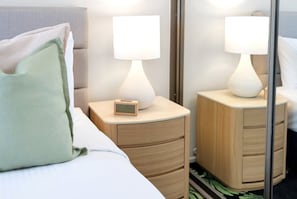 A hotel room with a bed, a nightstand with a lamp, a small clock, and a mirror reflecting the lamp. - Belle Escapes Aquarius' Panoramic Jewel 11th Floor (Cairns City)
