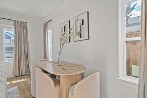 Ein Essbereich mit einem Holztisch, Stühlen und einer Vase mit Blumen. An der Wand befinden sich gerahmte Bilder und ein Fenster mit Vorhängen.