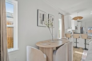 Een moderne eetruimte met een houten tafel, stoelen en een vaas met bloemen. Er hangen kunstwerken in de muur, een keuken op de achtergrond en een uitzicht op een houten terras door het raam.