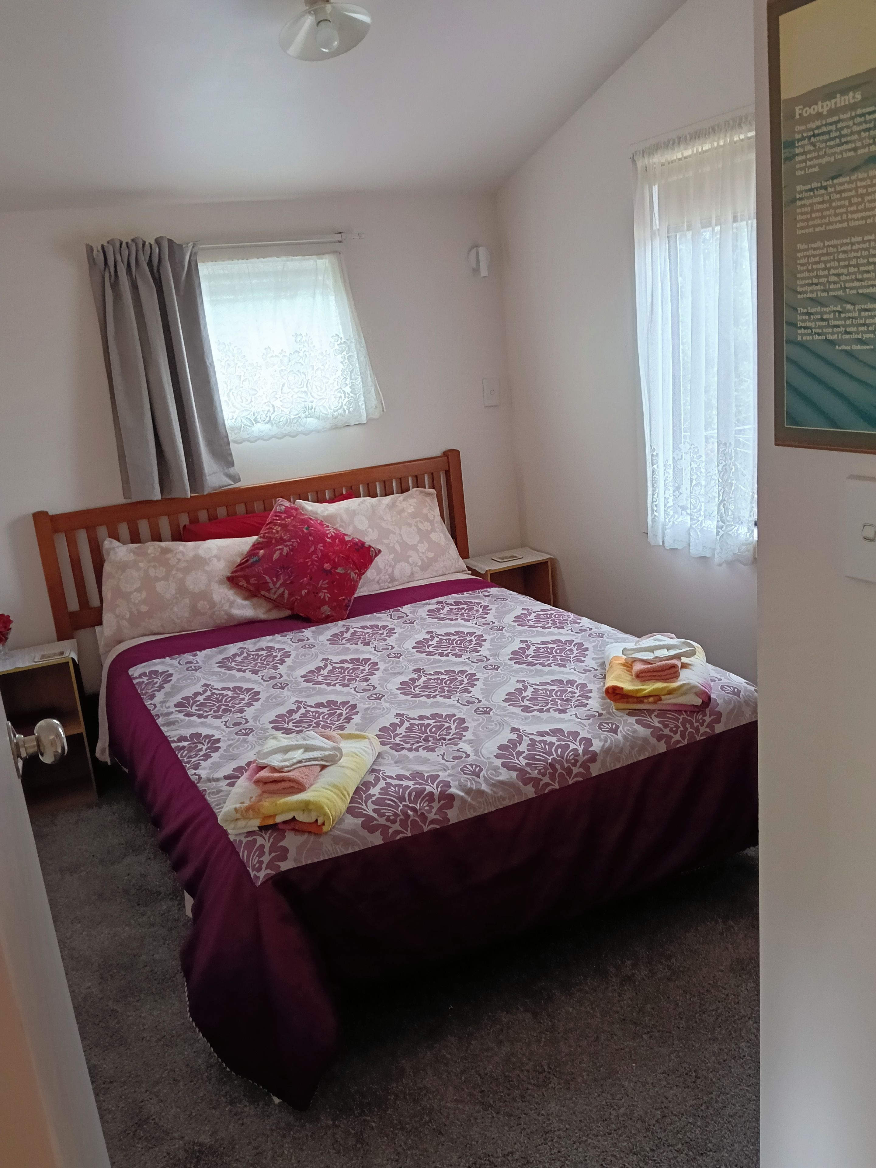 A bedroom with a wooden bed, a nightstand, a window with curtains, and a wall-mounted light fixture.