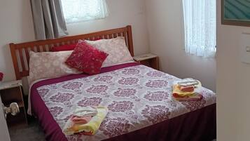 A bedroom with a wooden bed, a nightstand, a window with curtains, and a wall-mounted light fixture.