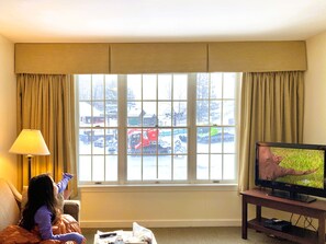 A hotel room with a large window, a television on a stand, and a child sitting on a couch.