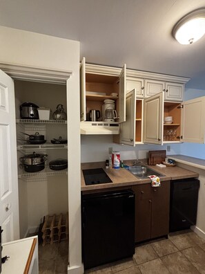 A compact kitchen with a dishwasher, a stove, and a sink. There are cabinets above the counter and a small pantry area with shelves.