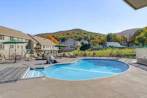 A swimming pool with lounge chairs and umbrellas, surrounded by buildings and a hilly landscape.