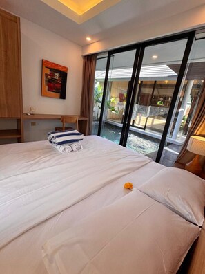 A hotel room with a large bed, a desk, a chair, a window with a view of a pool, and a painting on the wall. - Villa Jia by Kelle, Ubud (Gianyar)