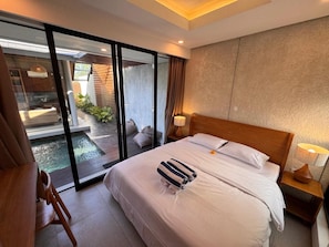 A hotel room with a bed, a wooden nightstand, a glass door leading to a pool area, and a view of the outdoors. - Villa Jia by Kelle, Ubud (Gianyar)