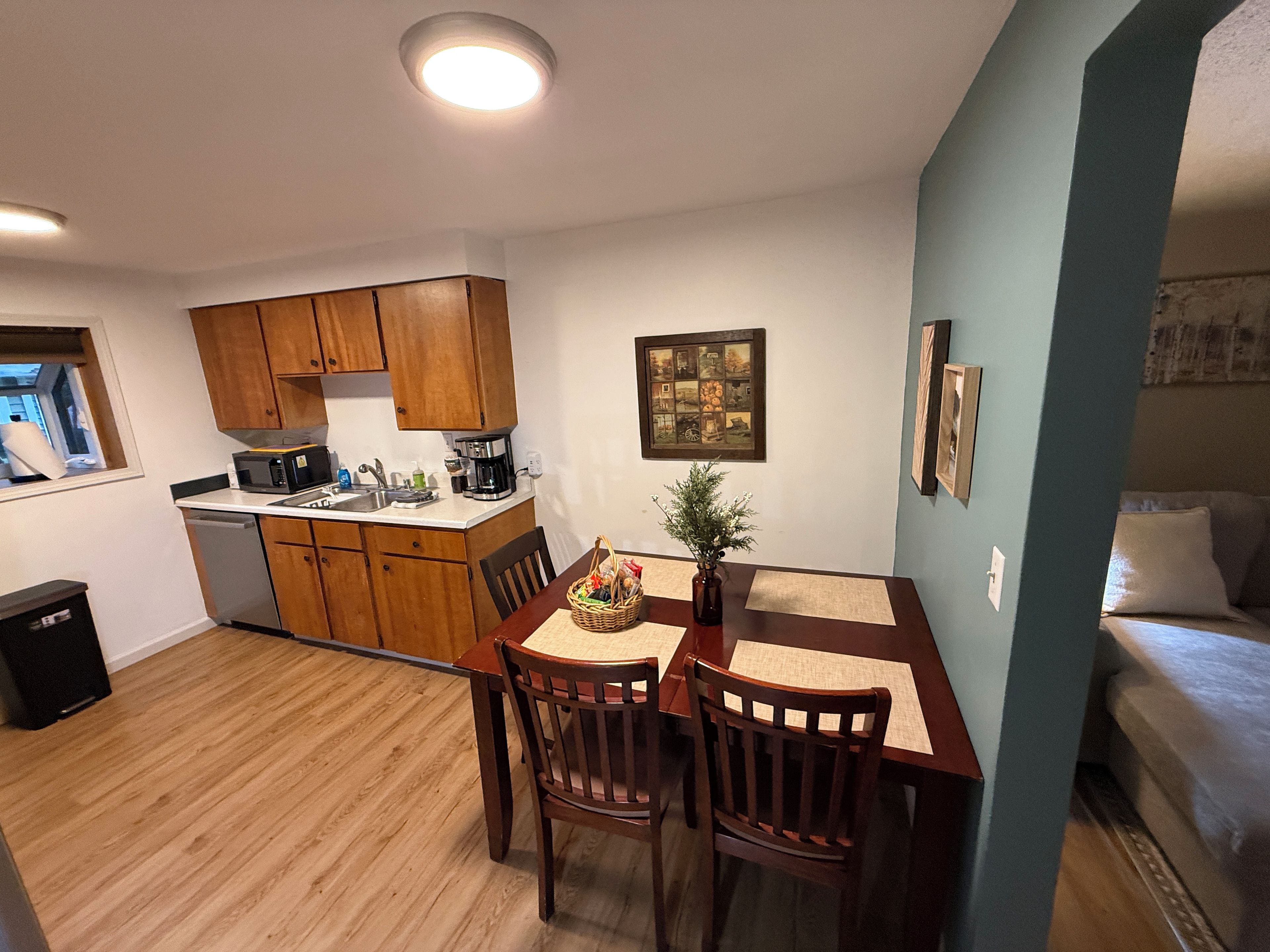 A compact kitchen with wooden cabinets, a dining table with chairs, a coffee maker, and a small potted plant.