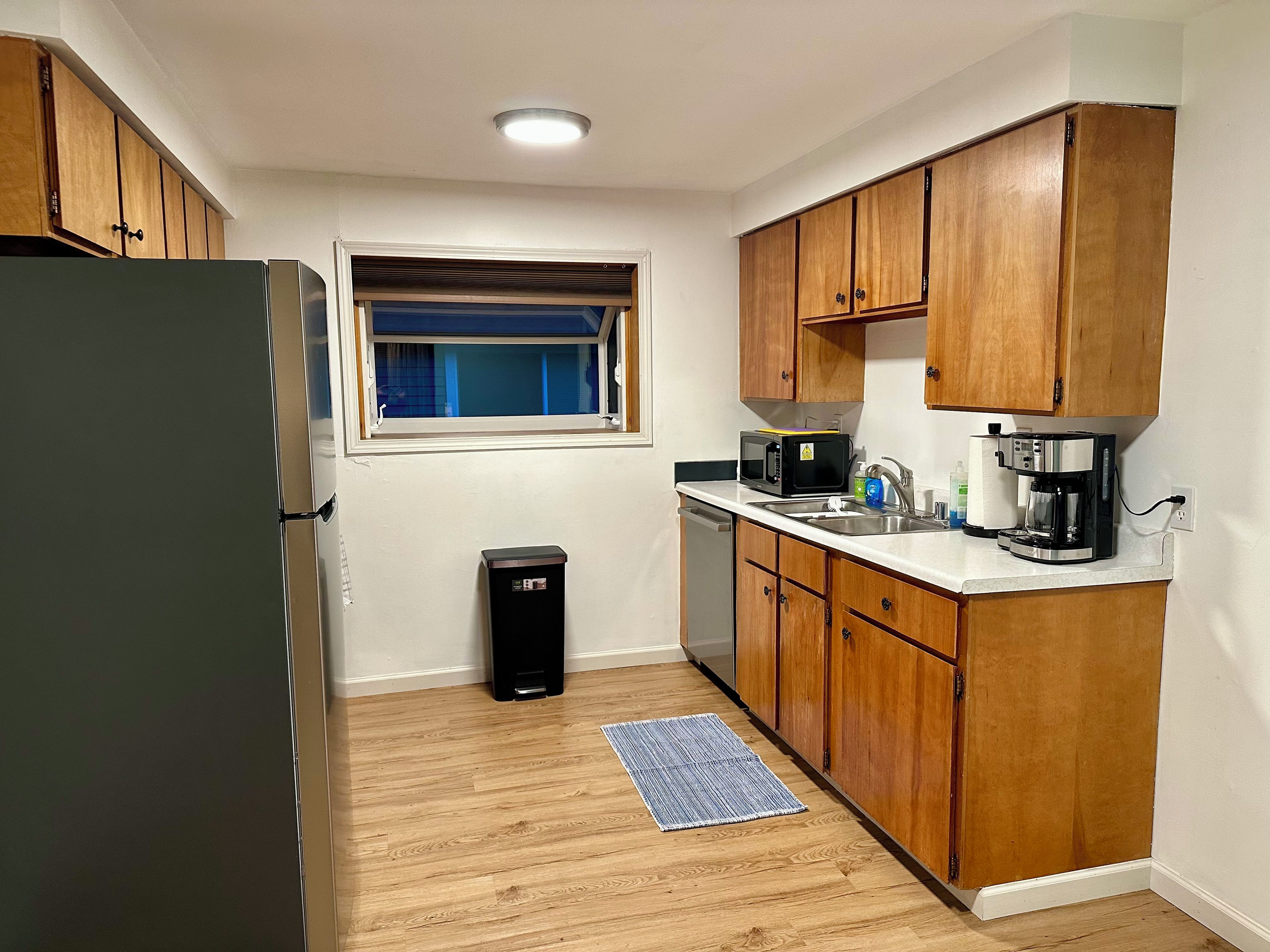 A compact kitchen with wooden cabinets, a stainless steel refrigerator, a microwave, a coffee maker, and a small trash bin.
