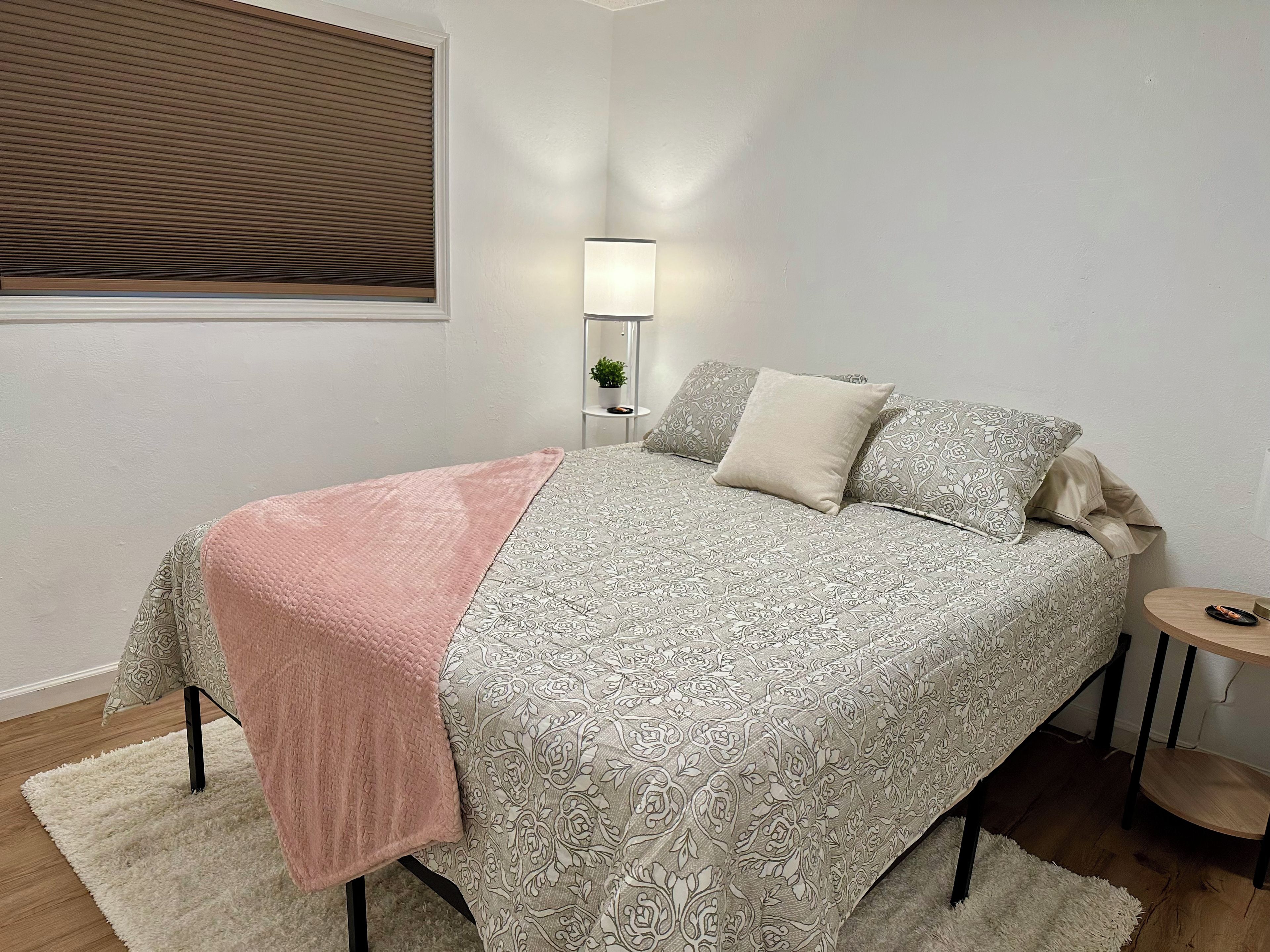 A bedroom with a bed, bedside table, lamp, and a window with blinds.