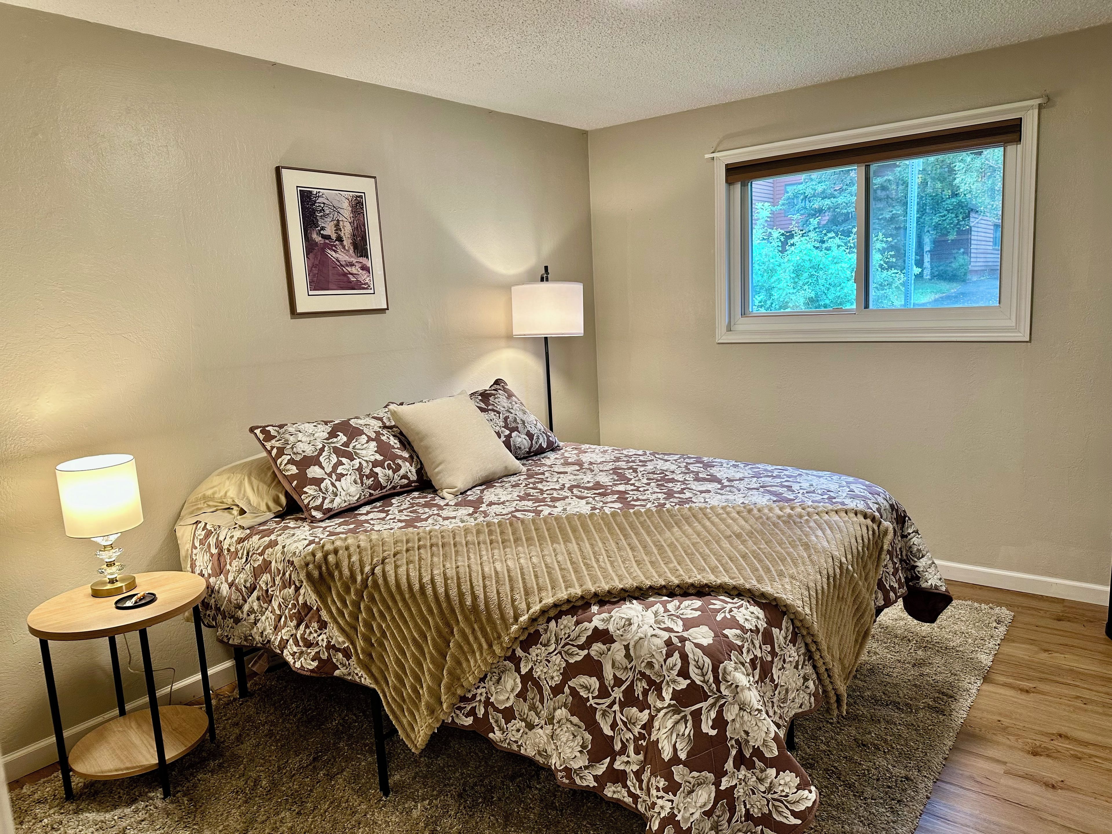 A bedroom with a bed, a bedside table with a lamp, a window, and a framed picture on the wall.