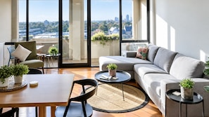 Ein modernes Wohnzimmer mit einer Couch, einem Esstisch und Stühlen. Auf dem Fensterbrett stehen Topfpflanzen und es bietet sich ein Blick auf die Stadtlandschaft.