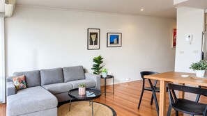 Ein modernes Wohnzimmer mit einem grauen Ecksofa, einem kleinen runden Couchtisch, einem Esstisch mit Stühlen und zwei gerahmten Bildern an der Wand.