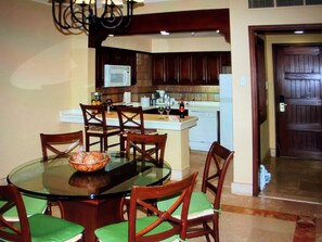 A dining area with a glass table surrounded by wooden chairs with green cushions, a chandelier, and a kitchen in the background. - 2BR/2B Presidential Suite at Pueblo Bonito Sunset with Exclusive Butler Service (Cabo San Lucas)