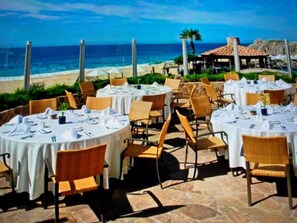 An outdoor dining area with round tables and chairs, overlooking a beach and ocean. - 2BR/2B Presidential Suite at Pueblo Bonito Sunset with Exclusive Butler Service (Cabo San Lucas)