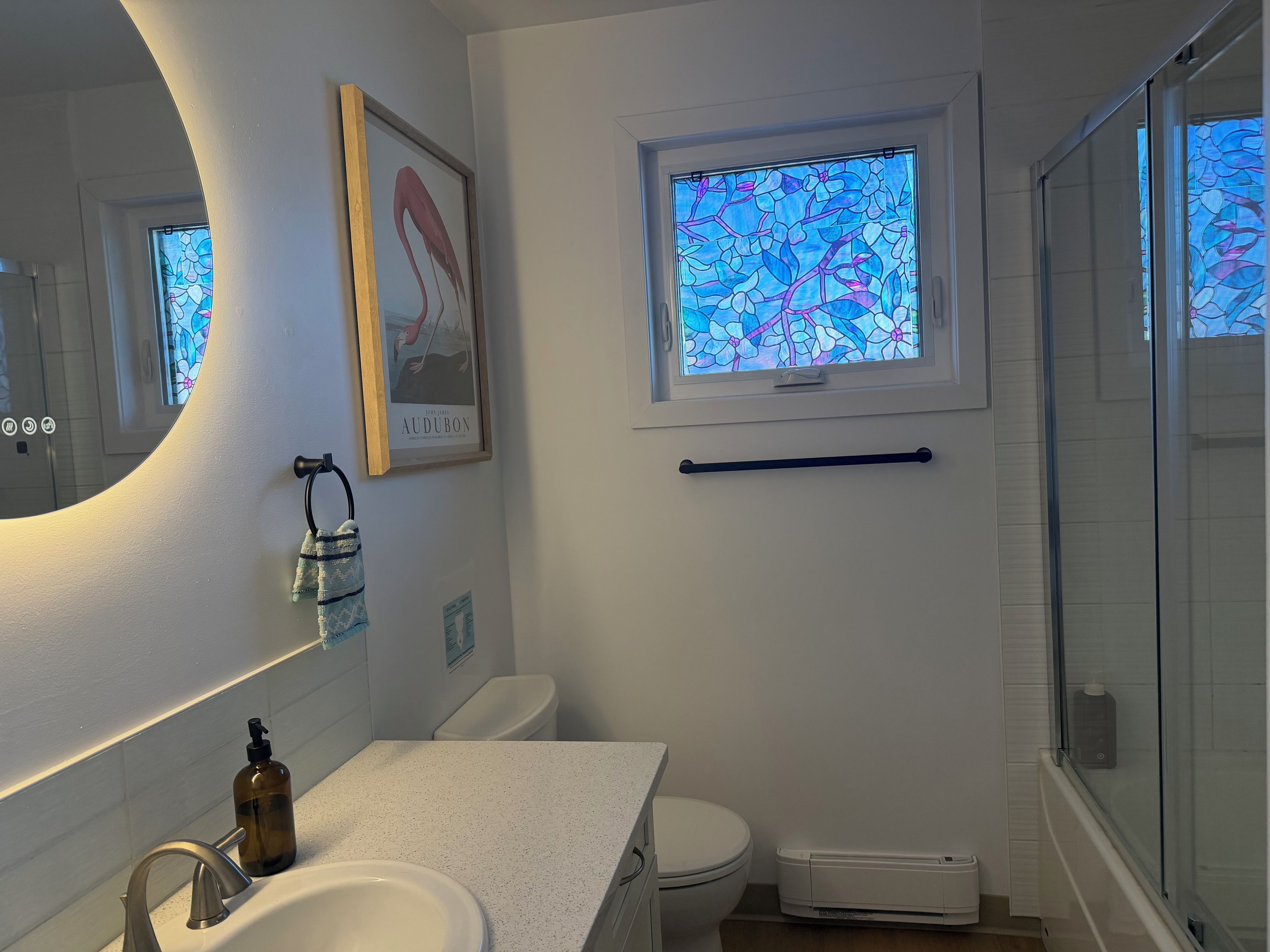 Un baño con un lavamanos blanco, un espejo, un cuadro enmarco, una ventana con vidrieras y una zona de ducha.