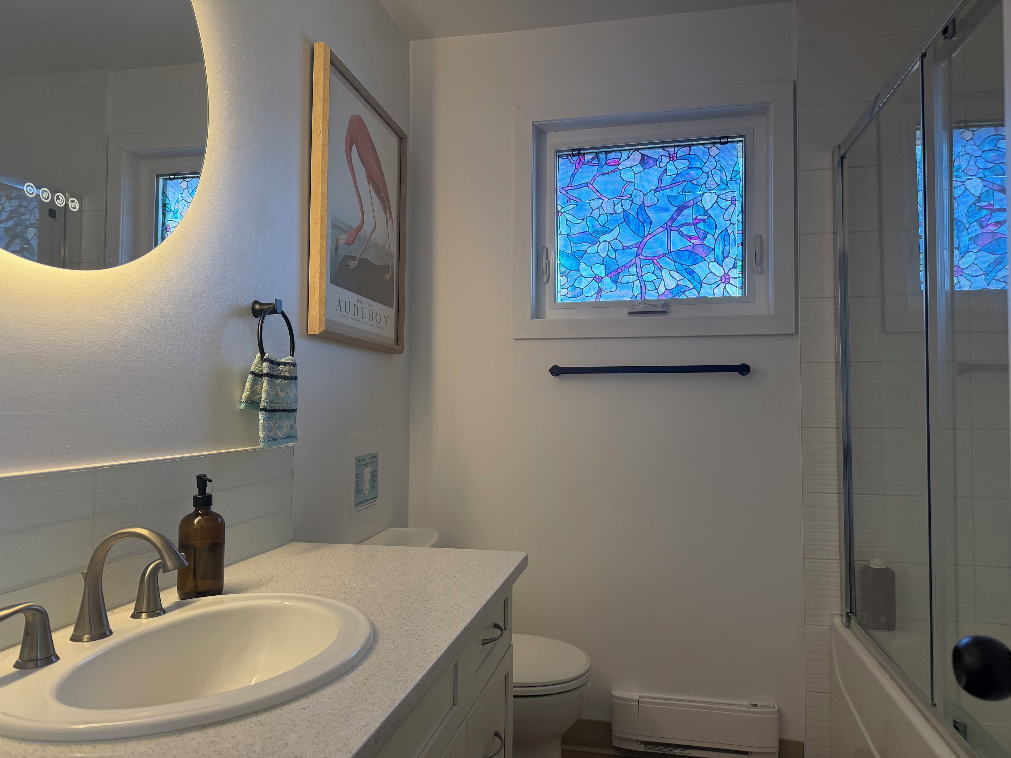 A bathroom with a white sink, a mirror, a framed picture, a window with a stained glass design, and a glass shower enclosure.