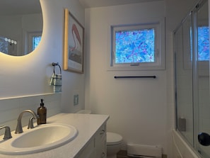 Un baño con un lavabo blanco, un espejo, un cuadro enmarco, una ventana con un diseño de vitral y una cabina de ducha de vidrio.