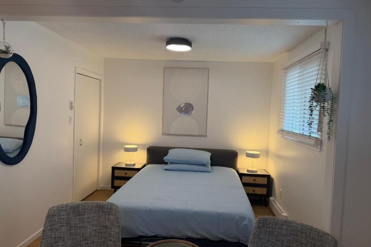 A bedroom with a bed, two bedside tables, a mirror, and a window with blinds.