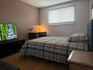 Un dormitorio con cama, mesita de noche, lámpara, televisor y ventana con persianas.