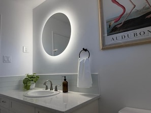 A modern bathroom with a round mirror, a sink, a towel rack, and a framed picture on the wall. - Holiday Collection (Winnipeg)