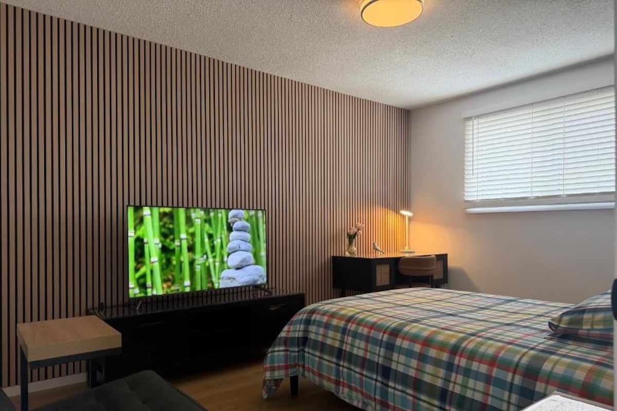 A bedroom with a television on a stand, a bed with a plaid bedspread, a bedside table, and a window with blinds.