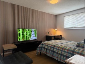 A bedroom with a television on a stand, a bed with a plaid bedspread, a bedside table, and a window with blinds. - Holiday Collection (Winnipeg)