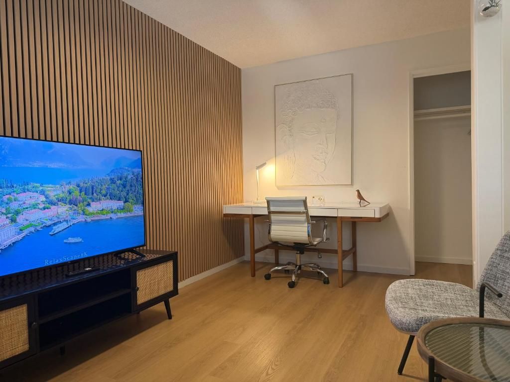 A modern living room with a flat-screen TV, a wooden slatted wall, a desk with a chair, and a glass table.