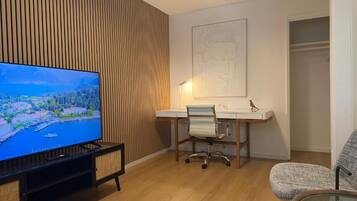 A modern living room with a flat-screen TV, a wooden slatted wall, a desk with a chair, and a glass table.