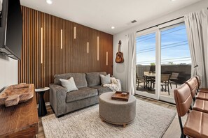 A modern living room with a grey sofa, wooden slat wall, a guitar, and a dining area with a table and chairs. - Modern Condo-5 Min to Broadway -Walk to Coffee (Nashville)